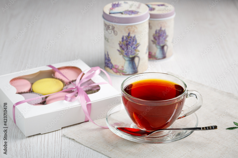 Multicolored macaroons in a box and a cup of tea on a light wood kitchen table. Morning tea and sweets. Multicolored macaroons in a box and a cup of tea on a light wood kitchen table. Morning tea and sweets.