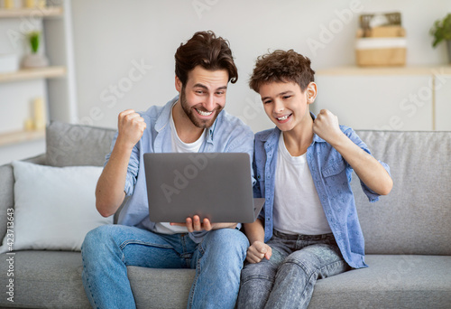 Internet tv. Overjoyed dad and his son watching sports online on laptop and cheering, raising hands with excitement