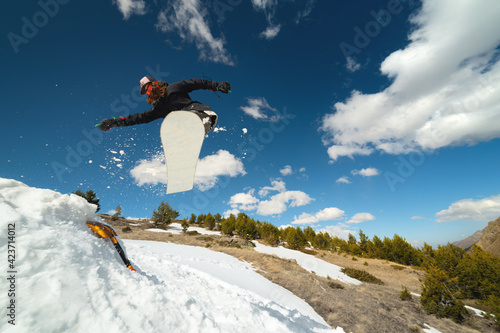 Snowboarder woman jumping from a kicker springboard from the snow on a sunny day in the mountains in a homemade snowboard park