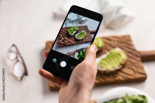 Hands take pictures on smartphone of two beautiful healthy sour cream and avocado sandwiches lying on board on the table. Social media and food concept