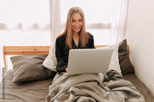 Smiling young business woman freelance student in domestic pajamas working remotely using technology and talking on a mobile phone looking at a laptop sitting in bed at home, selective focus