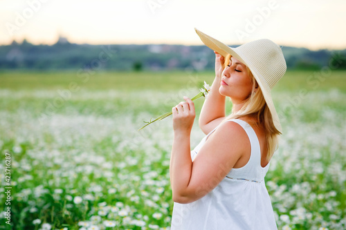 a beautiful plus-size girl in a hat and a white summer dress poses outdoors with camomiles. A plump girl in a meadow with daisies.