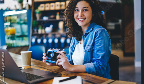 Portrait of successful hipster girl in trendy outfit holding vintage technology and smiling at camera, happy millennial woman with laptop computer and retro equipment spending time in coffee shop