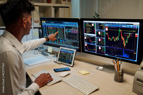 Professional young Indian trader working in dark office and pointing at computer screen with stock market information