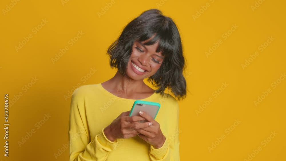 Young happy dark skinned Asian woman checks message on smartphone reads comments under post on webpage dressed in casual yellow jumper poses against orange background checks information in internet Young happy dark skinned Asian woman checks message on smartphone reads comments under post on webpage dressed in casual yellow jumper poses against orange background checks information in internet