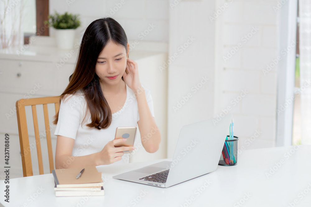 Young asian woman working laptop computer and reading smartphone on internet online on desk at home, freelance girl smiling using phone with social media, business and communication concept.