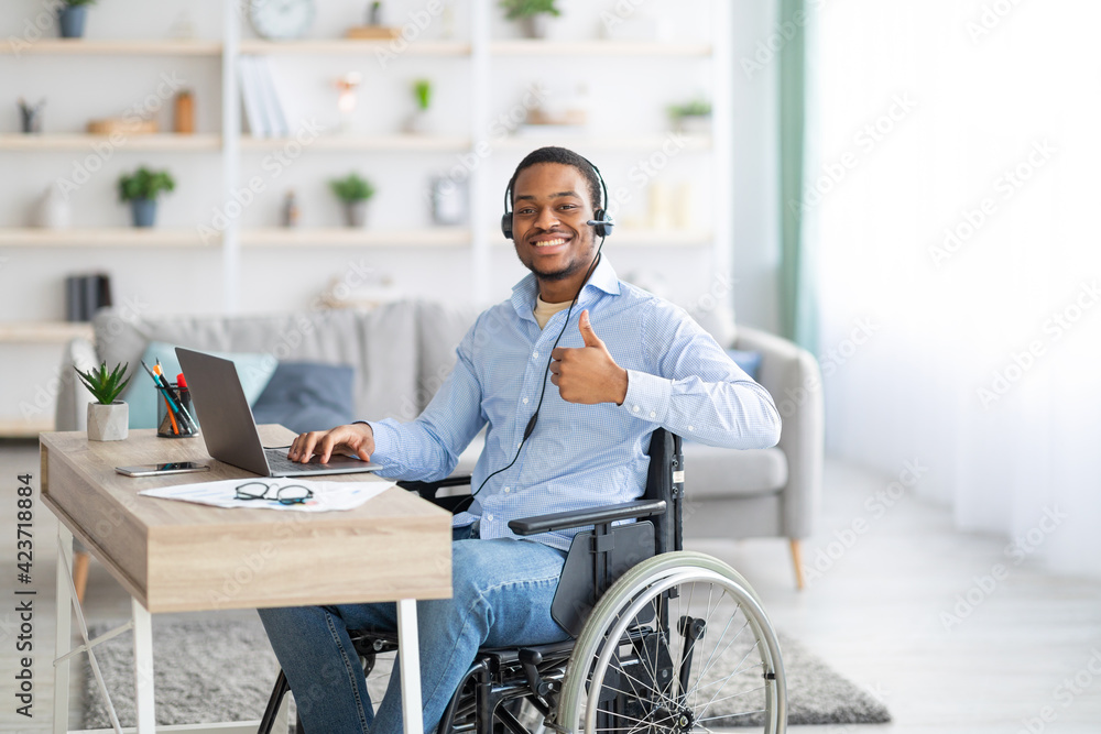 Positive disabled black man in headset using laptop computer for online work, showing thumb up gesture at home Positive disabled black man in headset using laptop computer for online work, showing thumb up gesture at home