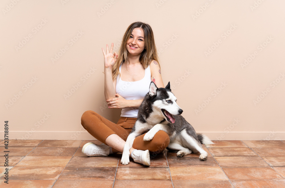 Young pretty woman with her husky dog sitting in the floor at indoors showing ok sign with fingers