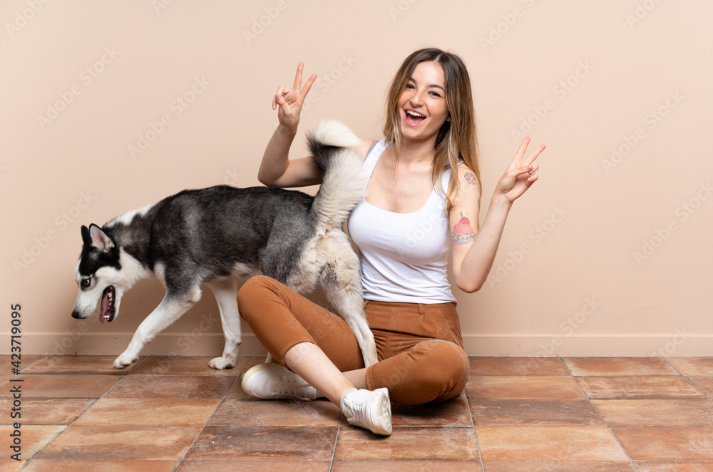 Young pretty woman with her husky dog sitting in the floor at indoors showing victory sign with both hands