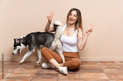 Young pretty woman with her husky dog sitting in the floor at indoors showing victory sign with both hands