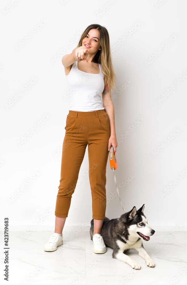 A full length young pretty woman with her dog points finger at you with a confident expression