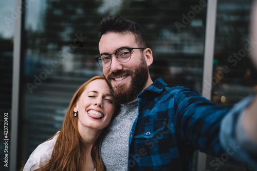 Selfie portrait of smiling female and male in classic eyewear making pictures during date in city, happy millennial hipster guys in casual clothing shooting influence vlog and posing at street