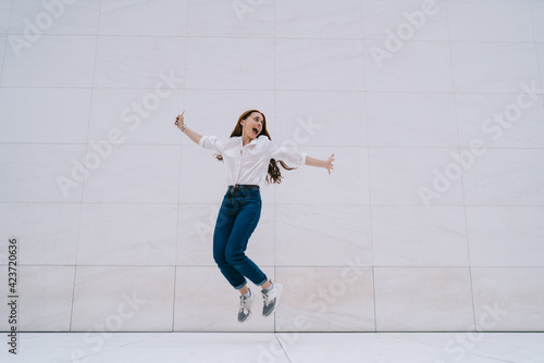 Excited hipster girl in casual outfit feeling satisfied happiness and freedom while jumping near promotional background with copy space area for fashion advertising, emotional female youngster