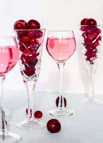 a glass of pink gin infused with cranberry among crystal glasses of berries on white background, cherry liquor or any red alcoholic cocktail closeup