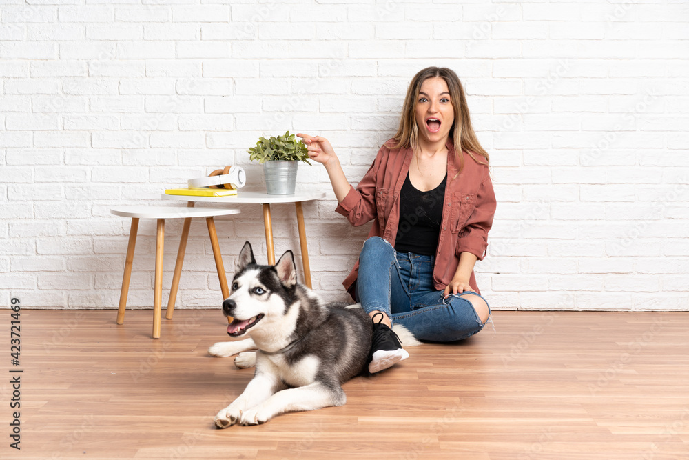 Young girl with her dog sitting in the floor at indoors surprised and pointing finger to the side Young girl with her dog sitting in the floor at indoors surprised and pointing finger to the side