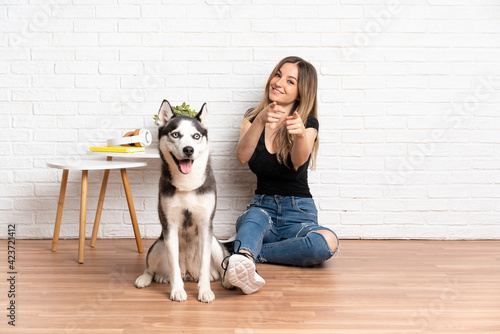 Young pretty woman with her husky dog sitting in the floor at indoors points finger at you with a confident expression