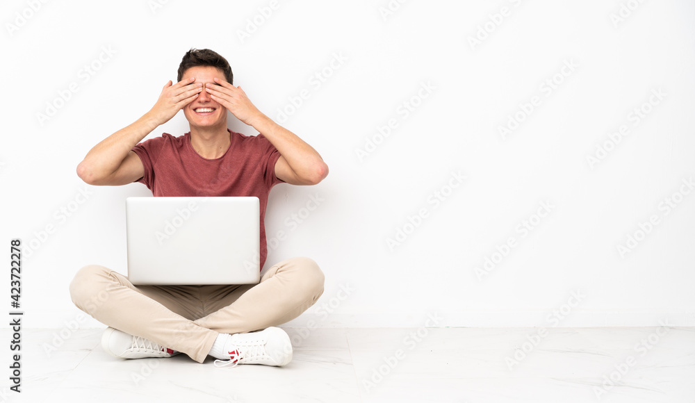 Teenager man sitting on the flor with his laptop covering eyes by hands