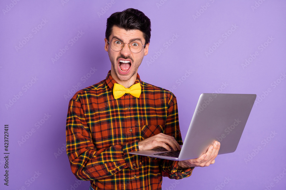 Photo of young crazy angry furious mad man in glasses having problem while working in laptop isolated on violet color background Photo of young crazy angry furious mad man in glasses having problem while working in laptop isolated on violet color background