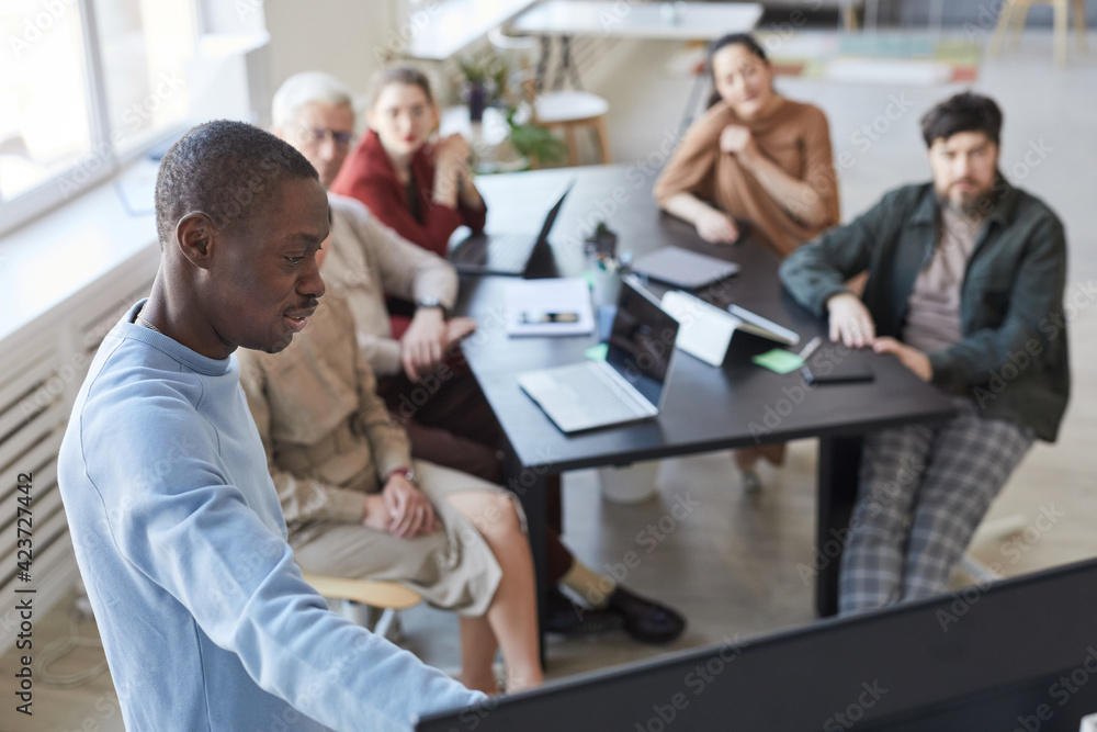 High angle portrait of African-American man giving presentation in office to diverse business team and pointing at digital screen, copy space High angle portrait of African-American man giving presentation in office to diverse business team and pointing at digital screen, copy space