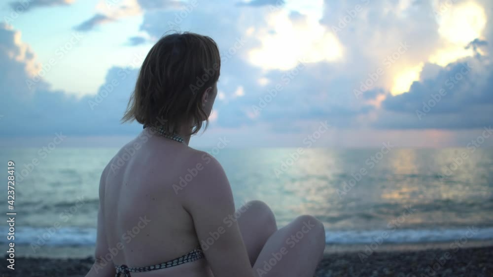 Middle-aged woman in swimsuit sits on pebble beach seashore admiring the sunset Middle-aged woman in swimsuit sits on pebble beach seashore admiring the sunset
