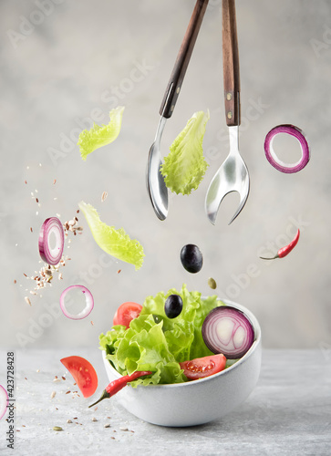 bowl with fresh salad, ingredients and cooking spoons in levitation on gray stone background