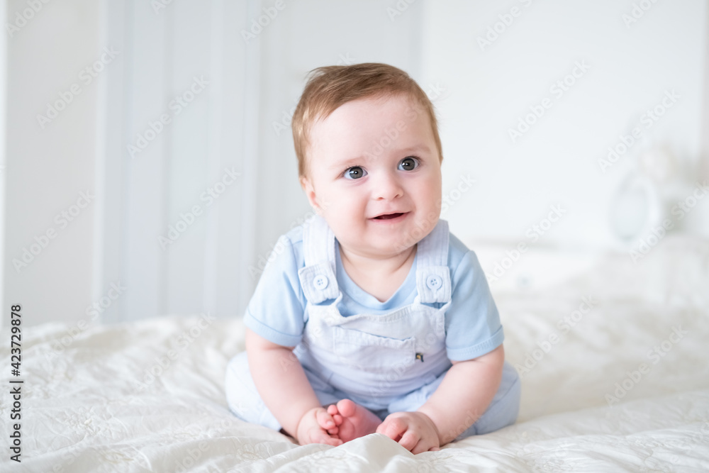 baby boy 6 months old in blu clothes smiling and sitting on white bed at home. baby boy 6 months old in blu clothes smiling and sitting on white bed at home.