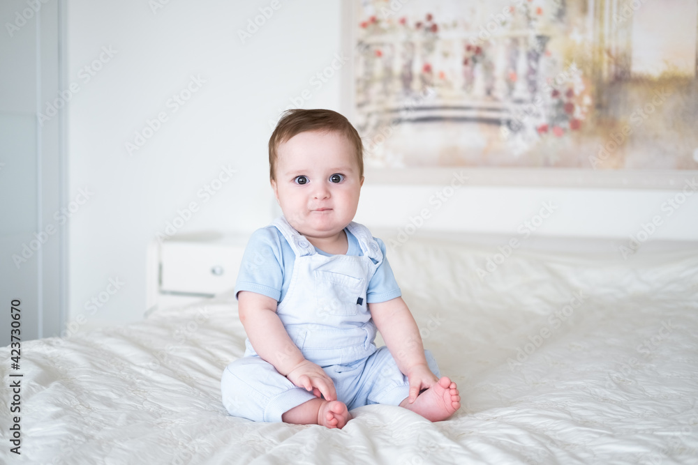 baby boy 6 months old in blu clothes smiling and sitting on white bed at home. baby boy 6 months old in blu clothes smiling and sitting on white bed at home.