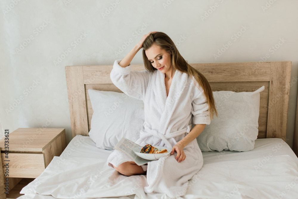 Young woman in white towel and robe in kitchen during quarantine. Hold donut on plate.