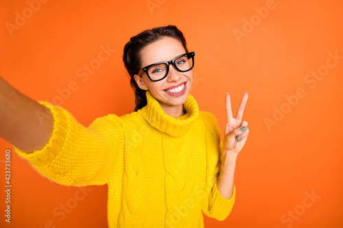 Photo of brown hair optimistic girl do selfie show v-sign wear spectacles yellow sweater isolated on orange color background