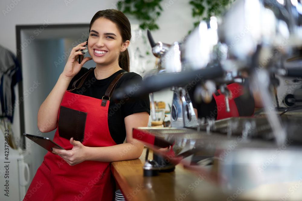 Smiling brunette woman waitress in uniform enjoying conversation on mobile phone making banking at work, prosperous female owner of coffee house calling to operator making order for retail business