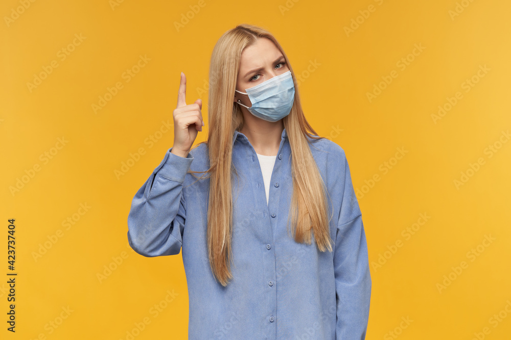 Nice looking woman, beautiful girl with long blond hair. Wearing blue shirt and medical face mask. People and emotion concept. pointing with finger upwards isolated over orange background Nice looking woman, beautiful girl with long blond hair. Wearing blue shirt and medical face mask. People and emotion concept. pointing with finger upwards isolated over orange background