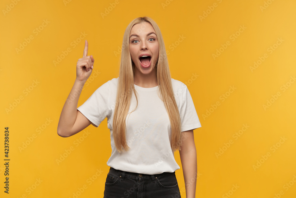 Portrait of attractive, adult girl with blond long hair. Wearing white t-shirt and black jeans. Keep mouth open. Watching at the camera and point with a finger upwards, isolated over orange background Portrait of attractive, adult girl with blond long hair. Wearing white t-shirt and black jeans. Keep mouth open. Watching at the camera and point with a finger upwards, isolated over orange background