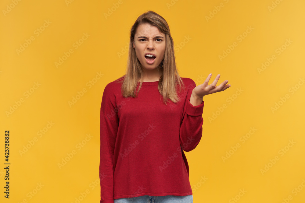 Shocked unsatisfied blonde girl looks discouraged, at a loss, with mouth opened, keeps one palm raised, wearing casual red sweater, standing isolated over yellow background Shocked unsatisfied blonde girl looks discouraged, at a loss, with mouth opened, keeps one palm raised, wearing casual red sweater, standing isolated over yellow background