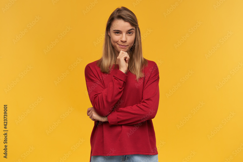 Tricky pretty young woman, blonde girl is plotting something. Wearing red sweater, keeps one hand near the chin. Thinking on some idea. Looking camera isolated over yellow background Tricky pretty young woman, blonde girl is plotting something. Wearing red sweater, keeps one hand near the chin. Thinking on some idea. Looking camera isolated over yellow background