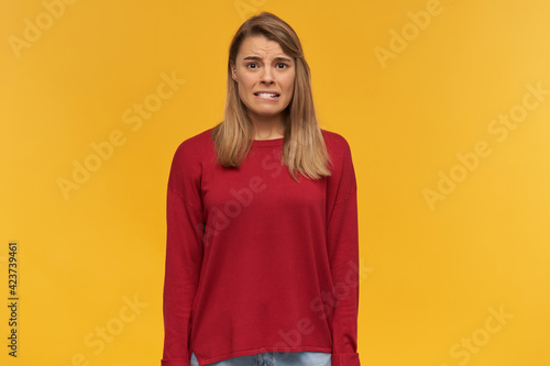 Girl looks looks with disgust, frowns, mouth opened, keeps mobile phone in her hand, black screen turned to the camera, points on it with index finger, isolated on a yellow studio background
