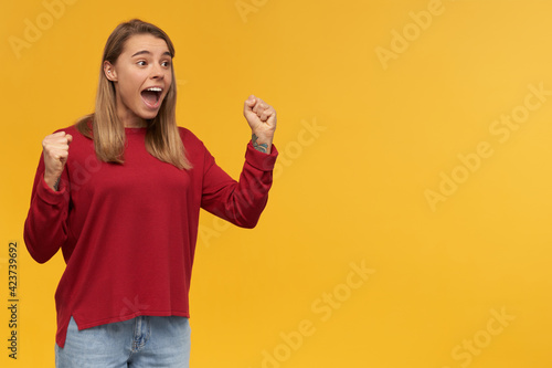 Surprised excited happily looking woman with blond hair. Wearing red sweater and glasses. Holding fists up celebrating her victory. Stand left half-side turned isolated over yellow background