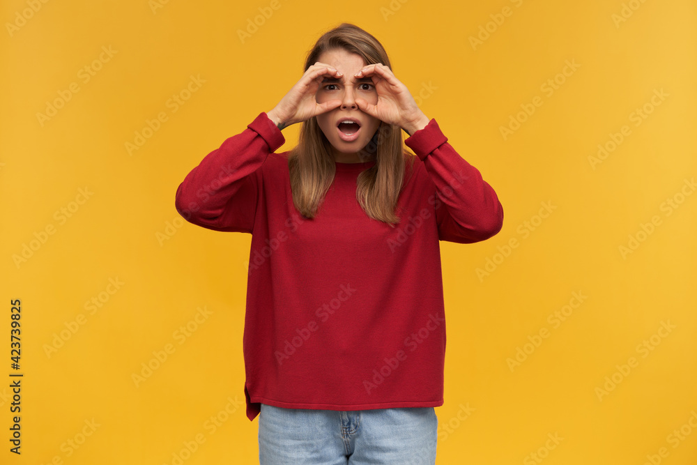indoor shot of excited young female starring into camera, wears red sweater and blue denim pants. standing over orange background with a fake binoculars. indoor shot of excited young female starring into camera, wears red sweater and blue denim pants. standing over orange background with a fake binoculars.
