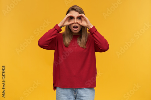 indoor shot of excited young female starring into camera, wears red sweater and blue denim pants. standing over orange background with a fake binoculars.