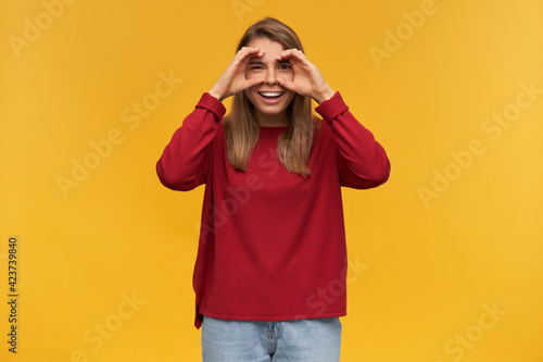 indoor shot of excited young female starring into camera, wears red sweater and blue denim pants. standing over orange background with a fake binoculars.