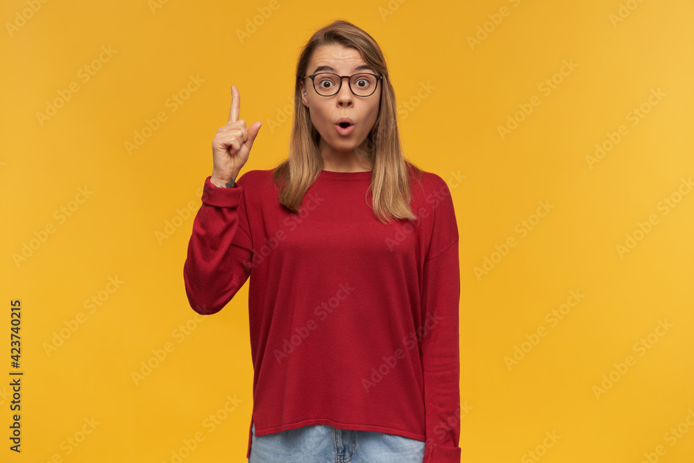 Young girl looking inspired, with rounded mouth, has blond hair. Wearing glasses. Pointing up with index finger on left hand like having a great idea. Looking camera isolated over yellow background Young girl looking inspired, with rounded mouth, has blond hair. Wearing glasses. Pointing up with index finger on left hand like having a great idea. Looking camera isolated over yellow background