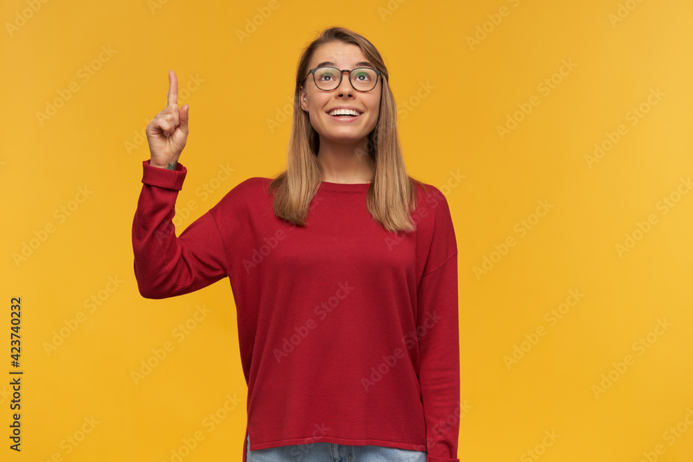 Excited looking woman, smiling girl with blond hair. Wearing red sweater and glasses. Pointing up with index finger on left hand like having a great idea. Looking up isolated over yellow background Excited looking woman, smiling girl with blond hair. Wearing red sweater and glasses. Pointing up with index finger on left hand like having a great idea. Looking up isolated over yellow background