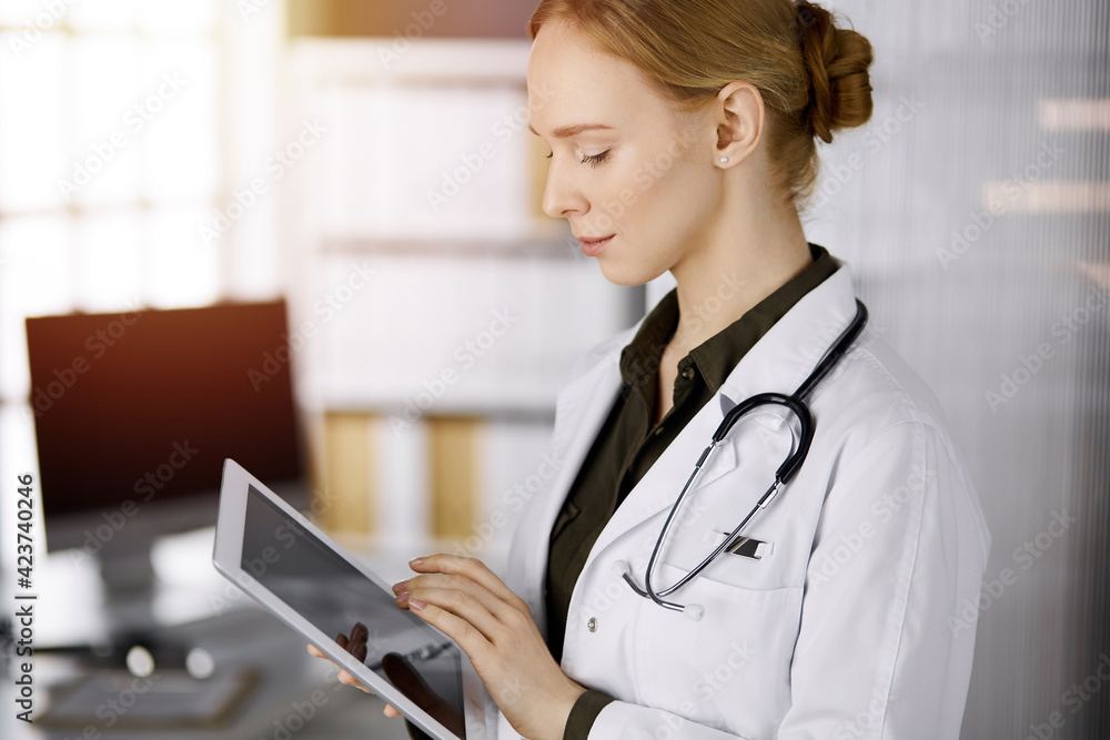 Cheerful smiling female doctor using tablet computer in sunny clinic. Portrait of friendly physician woman at work. Perfect medical service in hospital. Medicine concept Cheerful smiling female doctor using tablet computer in sunny clinic. Portrait of friendly physician woman at work. Perfect medical service in hospital. Medicine concept