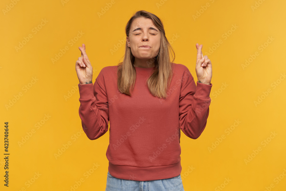 indoor shot of young student with crossed fingers keeps her eyes closed, biting her lip and thinking and praying about good result of her exams in high school