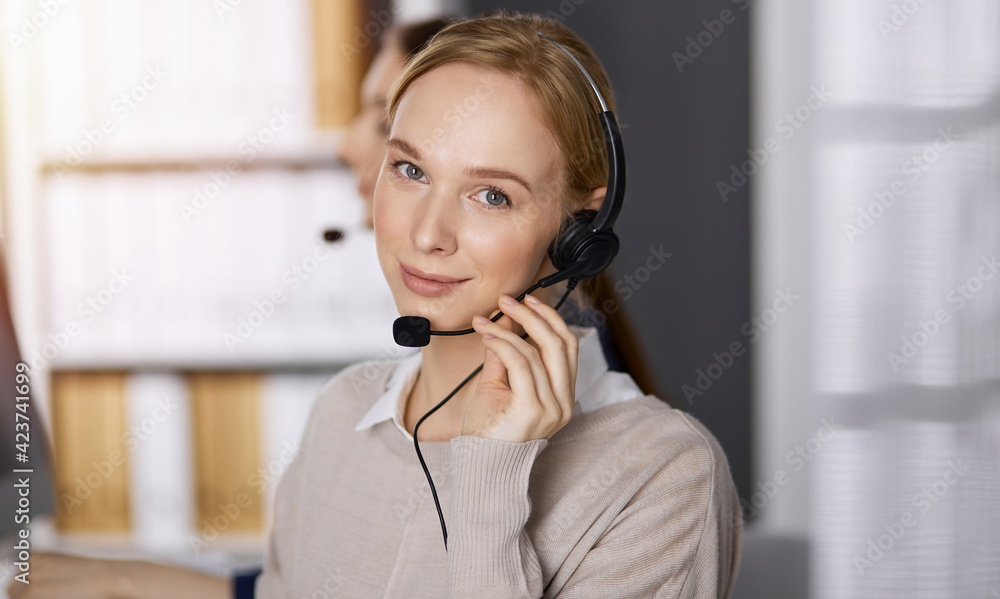 Businesswoman talking by headset while sitting in sunny office. Call center and diverse people group in business Businesswoman talking by headset while sitting in sunny office. Call center and diverse people group in business