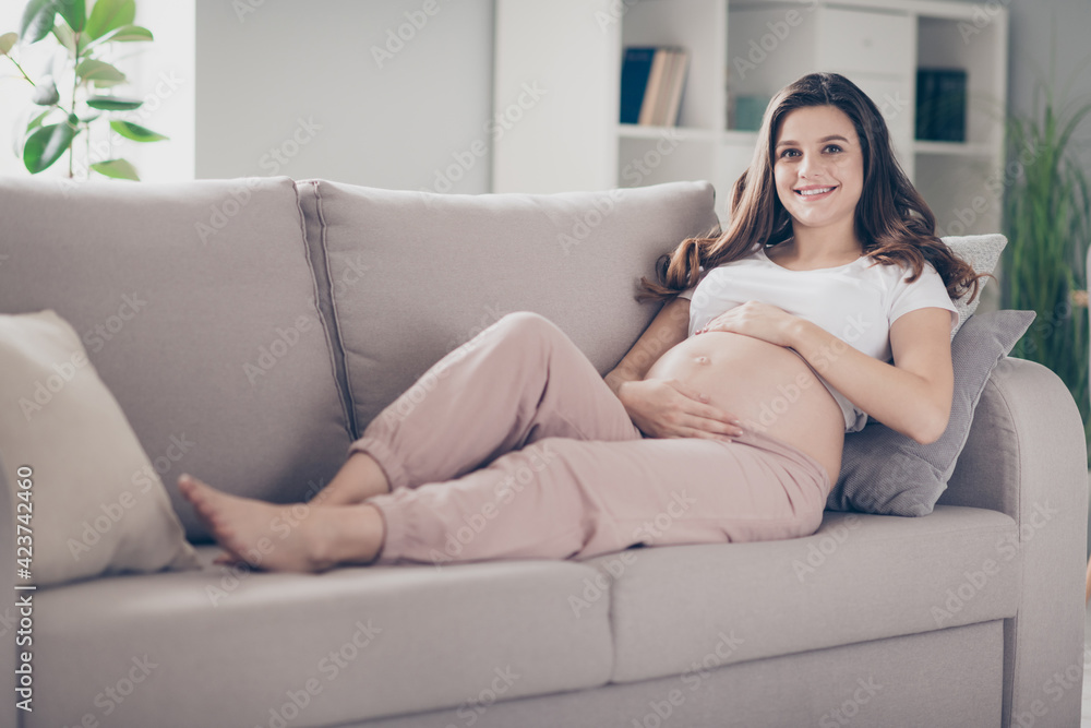 Full length photo of pretty happy young woman mother touch belly lie sofa in living room indoors inside house
