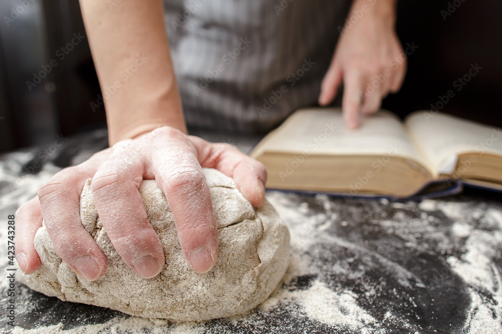 Baker holds one hand on the dough and the other on the cookbook, on the table sprinkled with flour. Bread making. Close-up.