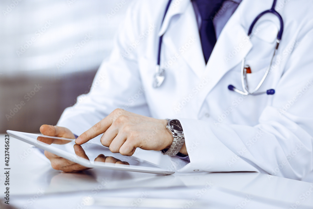 Unknown male doctor sitting and working with tablet computer in clinic at his working place, close-up. Young physician at work. Perfect medical service, medicine concept Unknown male doctor sitting and working with tablet computer in clinic at his working place, close-up. Young physician at work. Perfect medical service, medicine concept