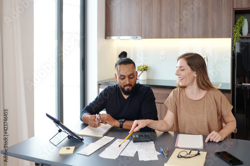 Smiling young woman pointing at bills on table and explaining boyfrend how to manage budget photo