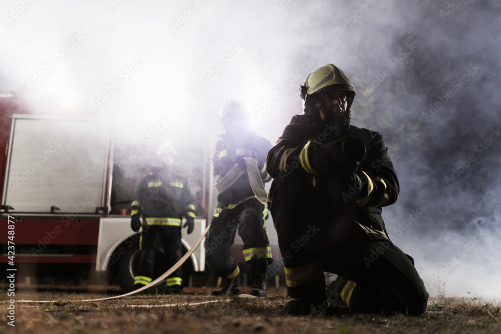 Naklejka premium Team of professional firefighters holding water hose in front of a firetruck with smoke in the air. Half silhouette.