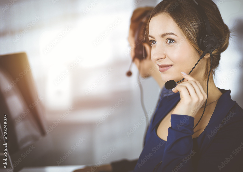 Two busineswomen have conversations with the clients by headsets, while sitting at the desk in a sunny modern office. Diverse people group in a call center. Telemarketing and customer service Two busineswomen have conversations with the clients by headsets, while sitting at the desk in a sunny modern office. Diverse people group in a call center. Telemarketing and customer service